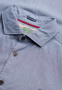 Chemise à rayures fines bleu et blanc, coupe régulière, col avec boutons visibles et étiquette verte « Superdry » à l'intérieur du col.