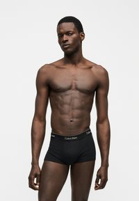 Μυώδες ανδρικό μοντέλο που φοράει μαύρα εσώρουχα Calvin Klein, στέκεται μπροστά σε ένα απλό λευκό φόντο, κοιτάζοντας ελαφρώς στο πλάι.