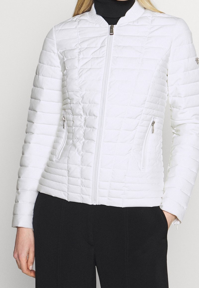Femme portant une veste matelassée blanche ajustée avec une fermeture éclair devant et des poches latérales zippées, associée à un pantalon noir et un col roulé noir.