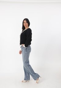 Cárdigan negro de punto con ribete blanco, combinado con jeans rectos de color azul claro. La modelo lleva sandalias de tacón alto blancas. Fondo blanco liso.