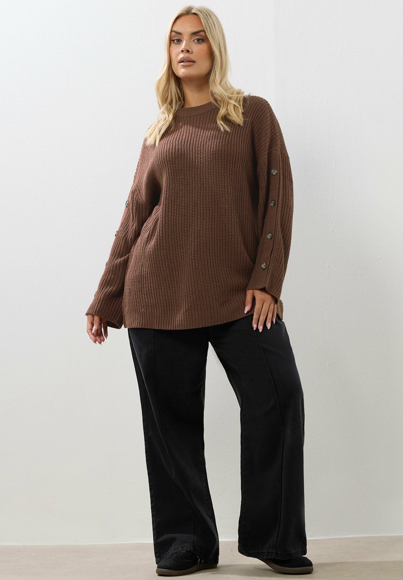 Brauner gerippter Strickpullover mit weiten Ärmeln, die mit Knöpfen an den Bündchen versehen sind, kombiniert mit schwarzen weitgeschnittenen Hosen und dunklen Schuhen.