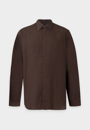 Homme debout portant une chemise marron à manches longues, un pantalon marron foncé et des baskets marron sur un fond clair uni.