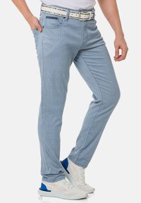 Helle blaue gestreifte Hose mit schmaler Passform, Fünf-Taschen-Design und einem weißen Gürtel mit dekorativen Nieten. Kombiniert mit weißen Sneakern.