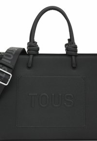 Czarna torebka o fakturowanej powierzchni z podwójnymi rączkami, odczepianym paskiem i wytłoczonym logo "TOUS" z przodu. Posiada węzłowe akcenty.