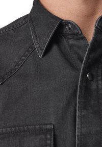 Camicia in denim nero con colletto a punta, bottoni a pressione e taschino. Il tessuto presenta una finitura testurizzata e dettagli di cucitura visibili.