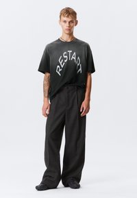 Črn grafični majica s kratkimi rokavi z besedo "RESTART" v srebrni barvi, upognjeni v lok, kombinirana z ohlapnimi sivo karirastimi hlačami in črnimi čevlji z teksturiranim podplatom.