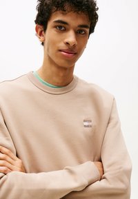 Jonge man met krullend, donkerhaar die een beige Tommy Jeans sweatshirt over een groene shirt draagt, met de armen gekruist, recht vooruit kijkend tegen een neutrale achtergrond.