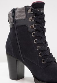 Kotníková bota z tmavě modrého semiše s masivním černým podpatkem, kovovými průchodkami a ozdobným šedým pleteným límcem. Má boční zip a propletené tkaničky.