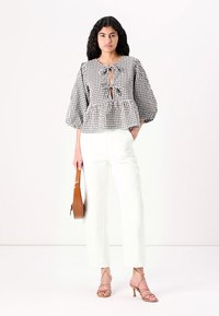 Blusa a scacchi bianco e nero con maniche a sbuffo, chiusura anteriore con nodo e vita a peplo, abbinata a pantaloni bianchi a vita alta e tacchi color tortora.