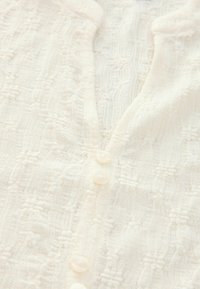 Gros plan d'un cardigan en maille texturée couleur crème avec un motif subtil et quatre petits boutons ronds sur le devant.