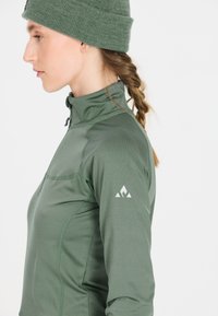 Kvinna i grön stickad mössa och grön jacka med kvartdragkedja och logotyp på ärmen, vänd åt vänster, med flätat hår bakom axeln mot vit bakgrund.