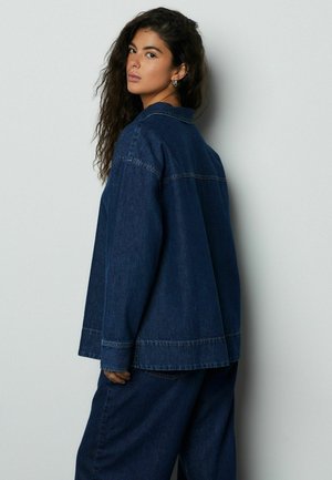 Žena s dlouhými kudrnatými vlasy, která má na sobě volnou tmavě modrou džínovou bundu a široké kalhoty v odpovídající barvě, dívající se přes rameno.