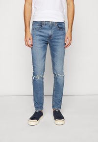 Vaqueros de mezclilla azul con rodillas desgastadas y corte estrecho. Combinados con una camiseta blanca y zapatillas deportivas azul marino. Textura suave y diseño clásico.