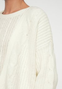 Κοντινό πλάνο ενός ατόμου που φοράει ένα λευκό πλεκτό πουλόβερ με υφή και διάφορα σχέδια πλεξίματος, με στρογγυλή λαιμόκοψη.