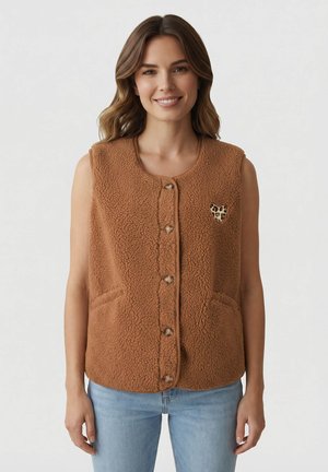 Donna sorridente che indossa un panciotto senza maniche in pile marrone con bottoni, patch a forma di cuore leopardato e jeans azzurri chiari, su sfondo neutro.