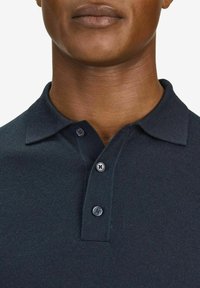 Tummansininen poolopaita, jossa on teksturoitu neulos, kauluksessa ja kahdessa napissa. Napit ovat pyöreitä, tummia ja kiiltävällä pinnalla.