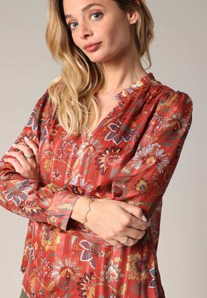 Blouse florale rouge avec un décolleté en V, des épaules froncées et des manches longues. Présente des motifs floraux complexes et un tissu léger et texturé.