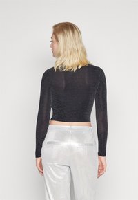 Musta pitkähihainen crop-top, jossa on kiiltävä tekstuuri, yhdistettynä hopeisiin korkeavyötäröisiin housuihin, joissa on takataskut ja sileä pinta.