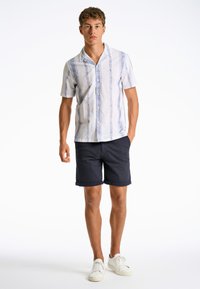 Camisa blanca de manga corta con patrones azules, combinada con pantalones cortos azul marino y zapatillas blancas. Diseño de cuello clásico y botón en la parte frontal.