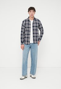 Καρό πουκάμισο σε ναυτικό μπλε και λευκό με κουμπιά, συνδυασμένο με ανοιχτόχρωμο μπλε τζιν. Το μοντέλο φοράει απλό λευκό μπλουζάκι από μέσα και αθλητικά παπούτσια.