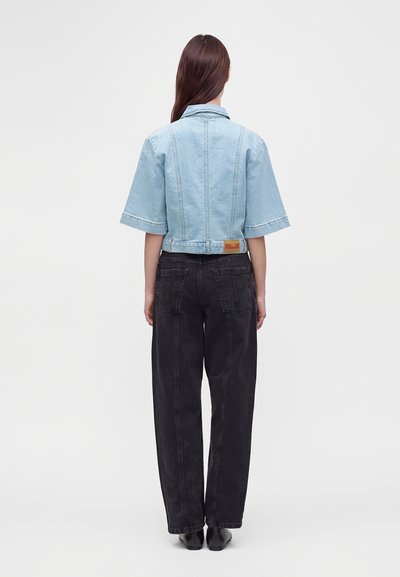 Personne aux longs cheveux bruns portant une veste en jean bleu clair à manches courtes et un jean noir ample, debout, dos tourné.