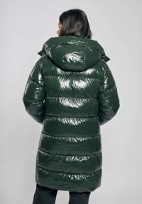 Kobieta widziana od tyłu, ubrana w błyszczącą, ciemnozieloną, puchową kurtkę z dużym kapturem oraz czarne spodnie na jednolitym tle.