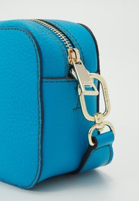 Tyrkysová kožená taška se zlatým zipem, doplněná odnímatelným zlatým klipem. Má prošívané okraje a obdélníkový tvar.