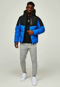 Padded jacket in black and bright blue, featuring a zip closure, hood, and sleek design, paired with grey joggers and white sneakers.