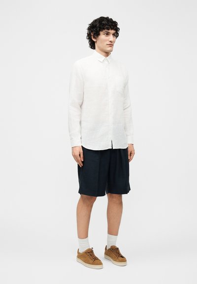 Νέος άνδρας με σγουρά σκούρα μαλλιά, φορώντας λευκό πουκάμισο με κουμπιά, μαύρα σορτς μέχρι το γόνατο, λευκές κάλτσες και καφέ αθλητικά παπούτσια, στέκεται μπροστά σε απλό φόντο.
