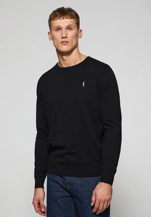 Suéter negro de punto con mangas largas y cuello redondo. Presenta un pequeño logotipo blanco en el lado izquierdo del pecho. Textura suave, corte clásico.