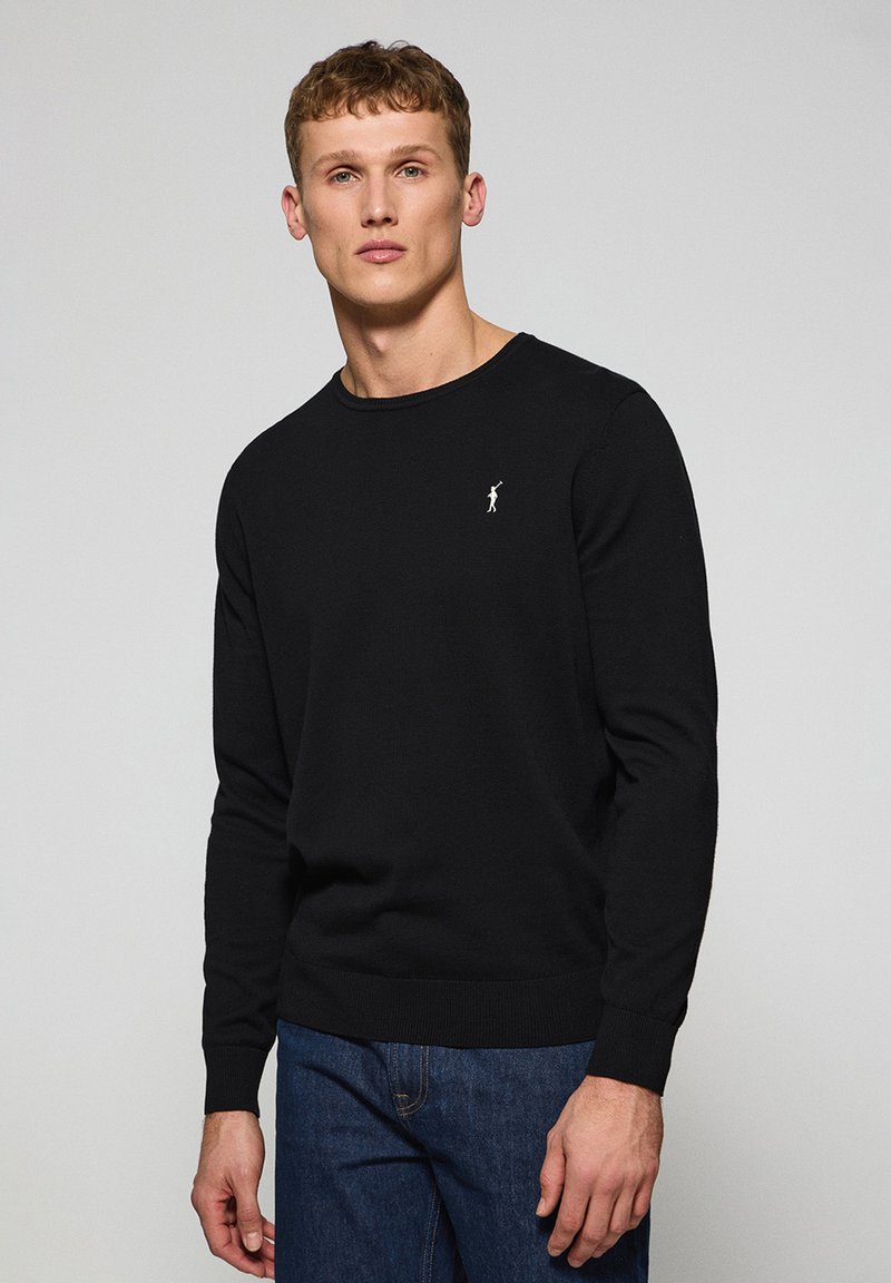 Suéter negro de punto con mangas largas y cuello redondo. Presenta un pequeño logotipo blanco en el lado izquierdo del pecho. Textura suave, corte clásico.
