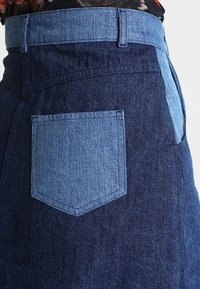 Jupe en denim dans deux nuances de bleu. Présente une taille haute, une ceinture contrastante en bleu clair et une poche rectangulaire au dos. Texture lisse.
