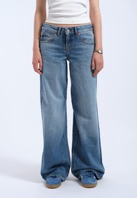 Jean en denim bleu clair à jambes larges avec une taille mi-haute, un léger effet délavé, et un design classique à cinq poches. Associé à des baskets bleues.
