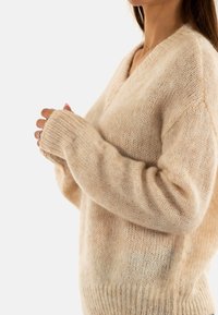 Suéter de lana beige con escote en V, corte holgado y puños acanalados. Textura suave y apariencia ligera con un acabado ligeramente peludo.