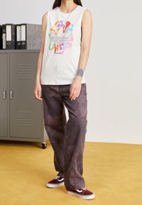 Wit mouwloos T-shirt met kleurrijk grafisch ontwerp en logo; gecombineerd met losse, donkerpaarse broek en bordeauxrode sneakers. Achtergrond met kluisjes.