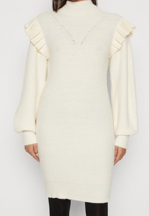Crèmekleurige gebreide jurk met gerimpelde schouders, lange ballonmouwen en een hoge hals, gedragen over zwarte legging.