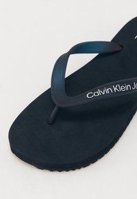 Tongs bleu marine avec une semelle d'accueil souple et une bride flexible. Présente un logo blanc "Calvin Klein Jeans" sur la bride.