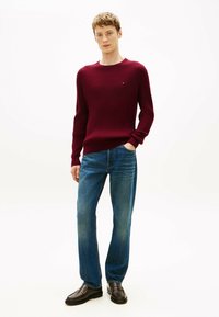 Pull côtelé bordeaux à manches longues, associé à un jean en denim bleu. Le mannequin porte des mocassins en cuir marron, debout devant un fond blanc uni.