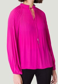 Blouse plissée fuchsia avec un col haut, détail en trou de serrure et manches ballon longues. Sa texture lisse et son tissu froncé améliorent son design.