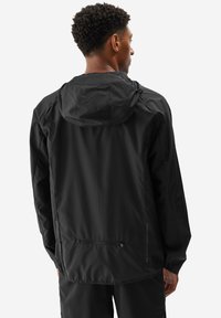 Mężczyzna odwrócony plecami, ubrany w czarną, lekką kurtkę z kapturem oraz pasujące czarne spodnie na jednolitym białym tle.