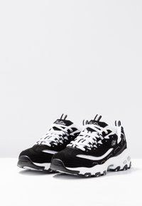 Black and white sneakers with a suede and mesh upper, thick white laces, cushioned sole, and prominent logo on the tongue and side.