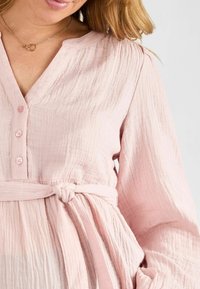 Abito rosa chiaro realizzato in tessuto testurizzato, con una chiusura frontale a bottoni, maniche lunghe e una cintura annodata in vita per un aggiustamento della forma.