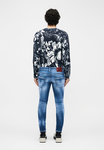 Άνδρας με σγουρά μαλλιά που φοράει μπλε πουλόβερ με λουλούδια, ξεθωριασμένο στενό τζιν και μαύρα παπούτσια με πλατφόρμα, στέκεται γυρισμένος από την πλάτη.