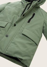 Jachetă de exterior verde, cu fermoar frontal, glugă, buzunare mari cu clapetă și accente negre. Textură netedă, dintr-un material rezistent la apă.