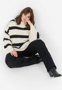 Übergroßer cremfarbener Pullover mit schwarzen Streifen, ausgestattet mit weiten Ärmeln und einem runden Ausschnitt. Kombiniert mit schwarzen Hosen und Stiefeletten.