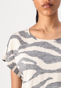 T-shirt à manches courtes en beige avec des rayures de zèbre grises, présentant un col rond et un tissu doux et texturé. Pas de fermetures visibles.
