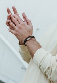 Mustmustehist kootud käevõru, millel on sile metallaktsent, kantud randmel. Käed on kokku pandud, nahatekstuur on näha.