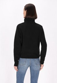 Fekete fleece pulóver magas gallérral, hosszú ujjak és laza szabás jellemzi, világoskék farmernadrággal párosítva. Simaság és minimalista dizájn jellemzi.