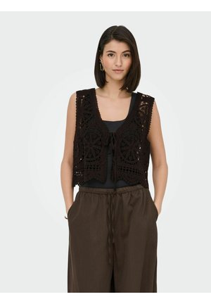 Donna che indossa un gilet nero all'uncinetto senza maniche sopra una maglia nera con pantaloni marroni larghi, mani nelle tasche, che guarda in avanti su sfondo semplice.