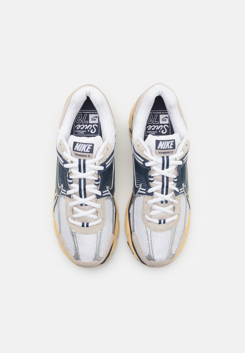 Zapatillas Nike Vomero 5 con una parte superior de malla blanca con detalles en marino y plata, suela de goma crema y cordones blancos.
